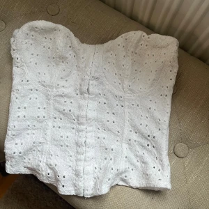 Vit broderad korsett-topp - Säljer en vit korsett-topp med broderade detaljer och små hålmönster. Toppen är axelbandslös och har en snygg formad byst. Materialet känns som bomull med ett fodrat insida för extra komfort. Perfekt för att skapa en chic och trendig look.