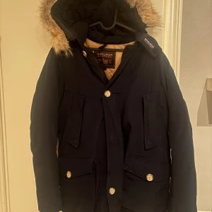 Svart woolrich arctic parka med päls - Parkas från Woolrich i svart med beige foder och huva med fluffig päls. Jackan har stora fickor med knappar och en rejäl huva som skyddar mot kyla. 