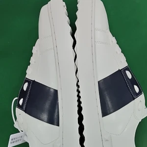 Valentino Garavani vita sneakers strl 43 - Snygga vita sneakers från Valentino Garavani med bred mörkblå rem över ovandelen och ikoniska nitar på sidorna och bak. Skorna har snörning, platt sula med mönstrad undersida och rund tå. Tillverkade i skinn med lyxig känsla och cool design.