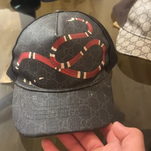 Gucci svart keps med röd orm - Svart Gucci keps med ikoniskt GG-mönster och en fet röd och vit orm på framsidan. Kepsen har mesh på sidorna och justerbar rem bak med klassisk Gucci-detalj. Materialet är polyester och syntet, perfekt för dig som vill sticka ut.