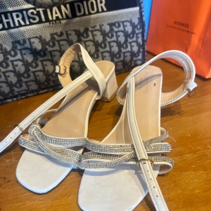 Vita sandaler med glittriga remmar - Säljer ett par vita sandaler från Mercury med fyrkantig tå och låg klack. Remmarna är dekorerade med glittrande stenar som ger extra bling. Justerbart spänne runt ankeln och platt sula för skön komfort. Perfekt för dig som gillar stilrena och glittriga detaljer.