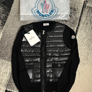 Moncler Cardigan  - Svart pufferjacka från Moncler med quiltad front och två fickor med knappstängning. Jackan har långärmade stickade partier på sidorna och Moncler-logga på ärmen. Dragkedja framtill och snygg, modern look som passar till streetwear.