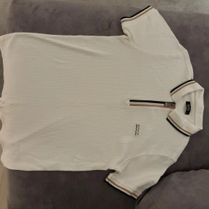 Vit pikétröja från Hugo Boss med zip - Säljer en fräsch vit pikétröja från Hugo Boss med ribbad struktur, halv dragkedja och snygga detaljer i svart och beige på krage och ärmslut. Boss-logga broderad på bröstet. Perfekt för en clean och stilren look.