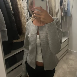 Grå quiltad cardigan jacka - Säljer en stilren grå cardigan jacka med quiltad struktur och dragkedja framtill. Jackan har rundad hals och lång ärm, perfekt för lager-på-lager och chill vardagsstil.