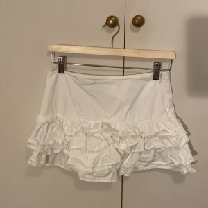 Vit volangkjol  - Supersöt vit kortkjol med flera lager volanger. Kjolen är gjord i mjuk bomull och har en luftig känsla. Perfekt för dig som gillar en feminin och lekfull stil. 