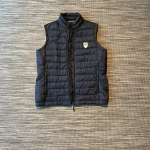 Svart  dunväst från Tuxer - Snygg svart dunväst från Tuxer med quiltad design och ståkrage. Västen har två praktiska fickor med dragkedja och en diskret logotyp på bröstet. Perfekt att slänga över en hoodie när det är kyligt ute.