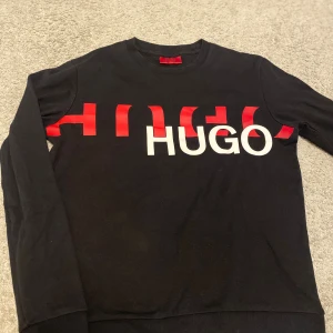 Svart långärmad tröja Hugo Boss - Svart långärmad tröja från Hugo Boss med stor HUGO-logga i vitt och rött över bröstet. Rund halsringning och mjukt bomullsmaterial som känns skönt mot huden. Perfekt för dig som gillar stilrena och moderna plagg med tydlig branding.