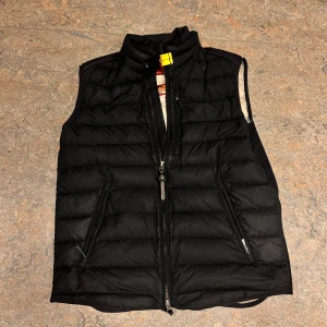 Svart dunväst från Parajumpers M - Svart quiltad dunväst från Parajumpers i super lightweight-modell. Västen har två fickor med dragkedja, hög krage och snygga detaljer på insidan i beige. Perfekt för lager-på-lager och har en stilren look med diskret logga på insidan.