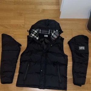 Svart dunväst från Burberry - Svart dunväst från Burberry med avtagbara ärmar och huva. Insidan har klassiskt rutigt Burberry-foder i grått, vitt och rött. Västen har två dragkedjefickor och en vit Burberry London England-logga på bröstet. Perfekt för att ge din outfit en lyxig touch.