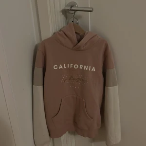 Rosa hoodie med tryck från H&M - Säljer en snygg hoodie från H&M i en soft rosa färg med texten CALIFORNIA framtill. Tröjan har huva, känguruficka och coola blockfärgade ärmar i beige och ljusgrått. Perfekt för chill dagar och streetstyle.