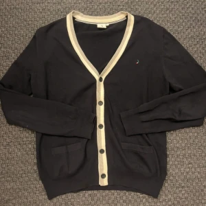 Marinblå kofta från Boomerang med beige kant - Marinblå kofta från Boomerang med beige kant längs v-ringningen och knäppningen. Klassisk design med två fickor framtill och diskret broderad logga på bröstet. Tillverkad i mjuk bomull och har långa ärmar. Perfekt för lager-på-lager-stil.