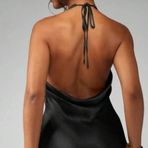 Svart halterneckklänning i satin - Snygg svart klänning i satin med halterneck och öppen rygg. Klänningen har en elegant drapering framtill och knyts i nacken. Perfekt för dig som vill ha en stilren och trendig look med lite edge.