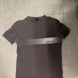 Grå BOSS t-shirt med silvertryck - Snygg grå t-shirt från Hugo Boss med ett coolt silvermönster och logga över bröstet. Klassisk rund hals och korta ärmar. Perfekt för dig som gillar stilrena plagg med en twist. Materialet är mjuk bomull som känns skönt mot huden.