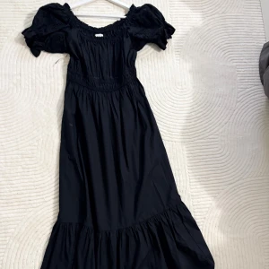 Svart långklänning med puffärm H&M - Svart långklänning från H&M med bred resår i midjan och puffiga kortärmar med broderade detaljer. Klänningen har en offshoulder-ringning och volang nedtill för extra flow. Perfekt för dig som gillar romantisk och trendig stil.