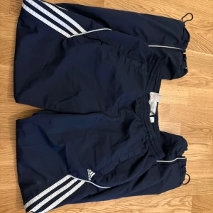Marinblå Adidas träningsshorts - Säljer ett par marinblå träningsshorts från Adidas med klassiska vita ränder på sidorna och diskreta reflexdetaljer. Shortsen har elastisk midja och är tillverkade i lätt polyester med meshfoder för extra komfort. Perfekta för sport eller chill dagar.