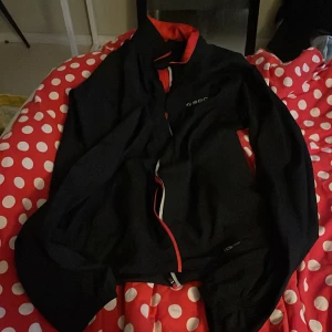 Svart vindjacka från SOC, strl 36 - Svart vindjacka från SOC med röda detaljer och meshfoder. Jackan har dragkedja framtill och står ut med sin sportiga look. Perfekt för blåsiga dagar och träning utomhus. TCS Wind-teknologi för extra skydd mot vind.perfekt som löparjacka med.