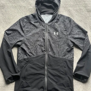 Svart vindjacka från Under Armour - Snygg svart vindjacka från Under Armour med huva och hel dragkedja. Jackan är som en tröja fast funkar som jacka när det är lite varmare. Inga skador. 