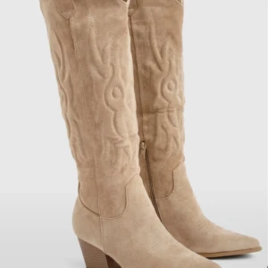 Beige cowboy boots i mocka - Snygga beige cowboy boots med spetsig tå och låg klack. Boots i mjuk mocka med dekorativa broderade detaljer längs skaftet och dragkedja på sidan. Perfekt för dig som vill ha western-vibe och stilren look. Ny pris 600kr