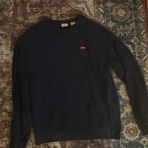 Mörkblå sweatshirt från Levi's - En stilren mörkblå sweatshirt från Levi's med klassisk rund hals och diskret röd logotyp på bröstet. Tröjan har långa ärmar och ribbade muddar vid ärmslut och nederkant. Perfekt för en avslappnad och snygg look.