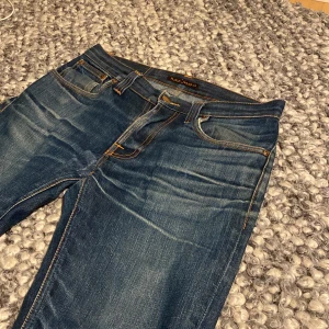Blå jeans från Nudie Jeans - Säljer ett par blå  jeans från Nudie Jeans i storlek 30/32. Jeansen är tillverkade i 100%  bomull och har klassiska orangea sömmar, samt snygga slitningar på hela byxan.