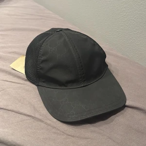 Svart keps från Gucci med mesh - Säljer en svart keps från Gucci med diskret GG-monogram över hela framsidan. Kepsen har böjd skärm, mesh på sidorna och justerbar snapback baktill. Tillverkad i polyester och bomull för en lätt och luftig känsla. Perfekt för dig som vill ha en stilren och lyxig accessoar. Den är lite sönder på backsidan men borde gå och fixa det, sällsynt och svår att få tag på