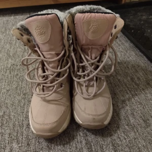 Beige vinterstövlar med fuskpäls - Säljer ett par beige vinterstövlar med snörning och mysig grå fuskpäls runt skaftet. Skorna är vattentäta och har en platt sula, perfekt för kalla dagar. Storlek 37.