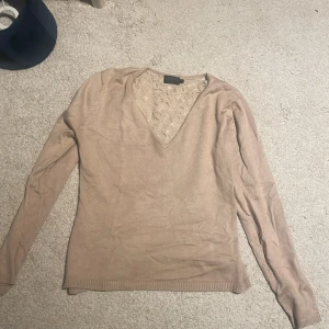 Beige v-ringad tröja med spetsrygg - Supersnygg beige tröja med v-ringning och lång ärm. Baksidan har ett genomskinligt spetsmönster med blommor som ger en unik och feminin touch. Perfekt för dig som vill ha något extra i garderoben!