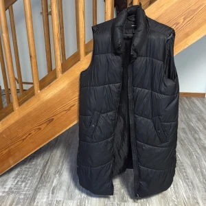 Svart lång dunväst från Vero Moda - Säljer en svart, quiltad dunväst från Vero Moda i storlek M. Västen är lång med hög krage, har två fickor med knappstängning och dragkedja framtill. Perfekt för lager-på-lager och snygg till både jeans och hoodies.