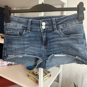 Blå jeansshorts från Nelly, strl 36 - Snygga blå jeansshorts från Nelly i storlek 36. Modellen har låg midja, fransiga kanter och coola vita broderade detaljer på bakfickorna. Klassisk femficksdesign med dubbla knappar framtill. Perfekt för sommarens alla äventyr!