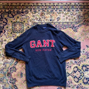 Mörkblå GANT tröja med röd logga M - Snygg mörkblå tröja från GANT med broderad röd och vit logga 'GANT NEW HAVEN' på bröstet. Tröjan har rund halsringning, ribbade muddar och är långärmad. Perfekt för dig som gillar klassisk college-stil.