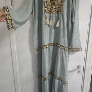 Ljusblå långklänning med broderi - Säljer en ljusblå långklänning från Al Walid med unikt guldigt broderi och dekorativa tofsar framtill. Klänningen har långa ärmar och detaljerade broderade band vid ärmslut, midja och nederkant. Materialet känns lätt och luftigt, perfekt för dig som gillar statement-plagg.