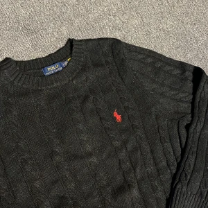 Svart stickad tröja från Polo Ralph Lauren - Svart kabelstickad tröja från Polo Ralph Lauren med rund halsringning och röd broderad logga på bröstet. Tröjan har långa ärmar och ribbade muddar vid ärmslut och nederkant. Perfekt för dig som gillar klassisk och stilren look.
