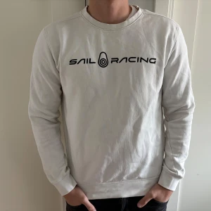 Vit Sail Racing sweatshirt - Vit sweatshirt från Sail Racing med svart logga tryckt på bröstet och en liten logga i nacken. Tröjan har rund halsringning, långa ärmar och ribbade muddar. Tillverkad i mjuk bomull för en skön och avslappnad stil.