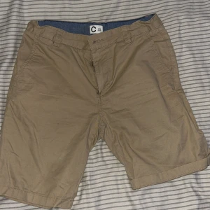 Beige shorts från C - Snygga beige shorts från Cubus i storlek 158, med klassisk passform och midwaist. De har fickor fram och bak, bälteshällor och stängs med knapp och dragkedja. Materialet är mjuk bomull, perfekt för varma dagar. Enkla att matcha med t-shirt eller hoodie. Jätte sköna att ha på sig överallt.