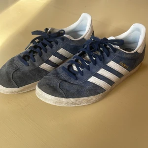 Adidas Gazelle blå/vita sneakers - Säljer ett par klassiska Adidas Gazelle sneakers i marinblå mocka med vita detaljer och snörning. Skorna har de ikoniska tre ränderna på sidan och guldtexten 'GAZELLE'. Perfekta för dig som gillar retrostil och streetwear.