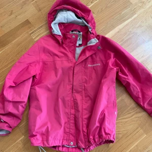 Rosa regnjacka från Didriksons - Snygg rosa regnjacka från Didriksons med huva och reflexdetalj på ryggen. Jackan har dragkedja framtill, justerbar nederkant och är helfodrad i ljusgrått. Perfekt för blöta dagar och håller dig torr med stil.