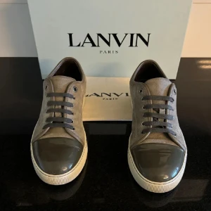 Grå sneakers från Lanvin med lackad tå - Snygga grå sneakers från Lanvin med ovandel i mocka och en glansig, lackad tå i mörkgrått. Skorna har vita sulor, grå snörning och diskreta sömdetaljer. Perfekta för dig som gillar stilrena och exklusiva sneakers med en twist.