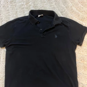 Svart pikétröja från U.S. Polo Assn - Klassisk svart pikétröja från U.S. Polo Assn med broderad logga på bröstet. Tröjan har korta ärmar, krage och två knappar framtill. Tillverkad i mjuk bomull som är skön mot huden och passar perfekt till jeans eller shorts.