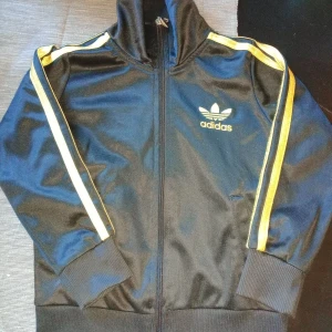Svart Barn Adidas track jacket med gula ränder - Storlek 98/104 Svart Adidas track jacket med gula ränder längs ärmarna och stort Adidas-tryck på ryggen. Klassisk modell med dragkedja framtill, ribbade muddar och hög krage. Materialet är glansig polyester som ger en sportig vibe. Perfekt för dig som gillar retrostil och streetwear.