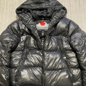 Svart pufferjacka från Zavetti Canada - Säljer en svart pufferjacka från Zavetti Canada med glansig finish, stor huva och feta dragkedjor. Jackan har broderad logga på ärmen och tryckt text på huvan. Insidan är fodrad med mjuk fleece för extra värme. Perfekt för kalla dagar.