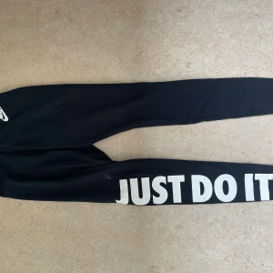 Svarta Nike leggings med tryck - Svarta leggings från Nike med stort vitt 'JUST DO IT.'-tryck längs ena benet och Nike-logga vid höften. Resår i midjan med textdetalj. Snygga och bekväma för träning eller chill. Gjorda i stretchigt syntetmaterial. Liten klorin fläck över texten ”just do it”