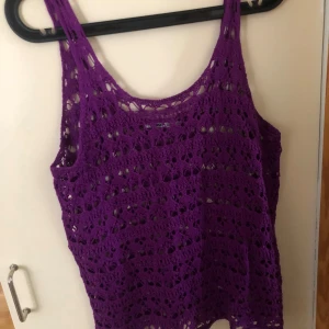 Lila virkad linnetopp - Säljer en lila virkad linnetopp med breda axelband och snyggt hålmönster. Perfekt för varma dagar och ger en cool, bohemisk vibe. Toppen är ärmlös och har en avslappnad passform som känns både trendig och bekväm.