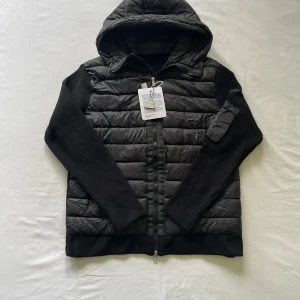 Moncler cardigan svart  - Svart Moncler cardigan med stickade ärmar och quiltad kropp. Jackan har huva, dragkedja framtill och en ficka med dragkedja på ena ärmen. Black label Moncler-logga på ärmen har NFC scan och coola detaljer. Perfekt för dig som vill ha en snygg look.