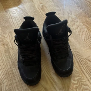 Svarta Nike Air Jordan 4 sneakers - Säljer ett par helsvarta Nike Air Jordan 4 sneakers med klassisk siluett och coola detaljer. Skorna har snörning, meshpaneler och den ikoniska Jumpman-loggan på plösen och hälen. Perfekta för dig som gillar streetwear och vill ha en stilren look.