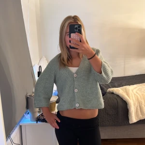 Ljusgrön stickad kofta från Lindex - Supermysig ljusgrön stickad kofta från Lindex med tre stora knappar framtill. Koftan är croppad och har trekvartsärmar, perfekt för lager-på-lager och chill vibes. Enkel att matcha med både jeans och kjol.