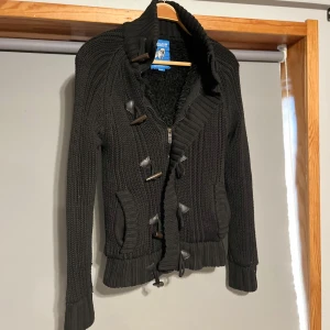 Fodrad cardigan  - Säljer en svart stickad cardigan med mysigt teddyfoder. Jackan har långa ärmar och stängs med både dragkedja och knappar. Perfekt för kyliga dagar!