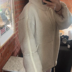 Beige teddyjacka med dragkedja - Mysig beige teddyjacka med hög krage och hel dragkedja framtill. Jackan har två fickor och en oversized passform, perfekt för lager-på-lager. Tillverkad i fluffigt fleece-material som håller dig varm under kyliga dagar.