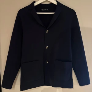 Mörkblå stickad cardigan från Zara - Snygg mörkblå stickad cardiganfrån Zara med klassisk krage och tre knappar framtill. Jackan har två stora fickor och är långärmad med en stilren och enkel design. Perfekt för dig som gillar en clean look och vill ha något som funkar till många outfits.