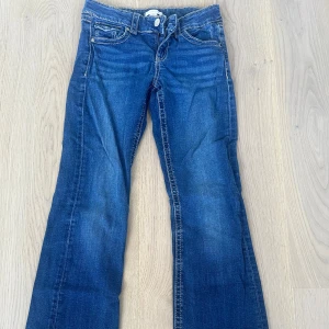 Blå bootcut jeans från Gina Tricot - Snygga blå jeans från Gina Tricot i storlek 158. Modellen har bootcut-ben, klassiska fem fickor och kontrastsömmar. Jeansen är i ett mjukt denimtyg och har knäppning med dragkedja och knapp framtill. Perfekta för dig som gillar en tidlös jeanslook.
