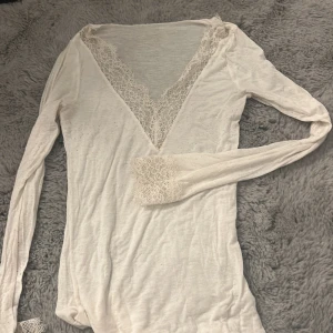 Vit långärmad topp med spetsdetaljer från intimissimi - Supersöt vit långärmad topp med djup v-ringning och spetsdetaljer både vid halsringningen och ärmsluten. Tunt och mjukt material som känns skönt mot huden. Perfekt för dig som gillar romantiska och feminina plagg.